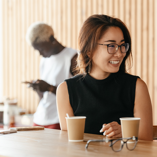 一个戴着眼镜的年轻亚洲妇女坐在咖啡馆里微笑着，后面是一个年轻的黑人妇女。