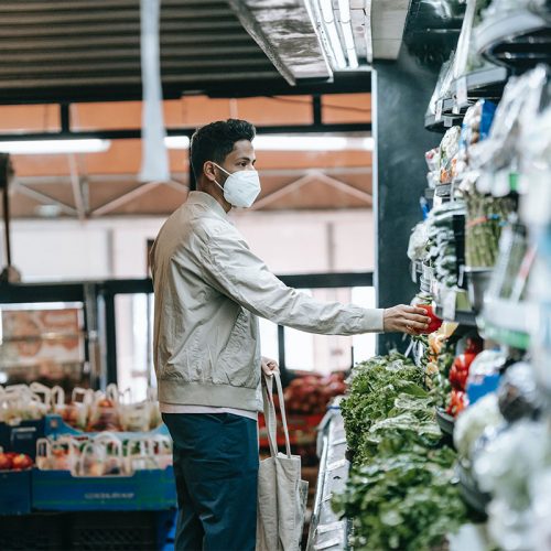 一名身穿奶油色夹克、蓝色裤子、戴着白色口罩的南亚男子从杂货店货架上拿起农产品