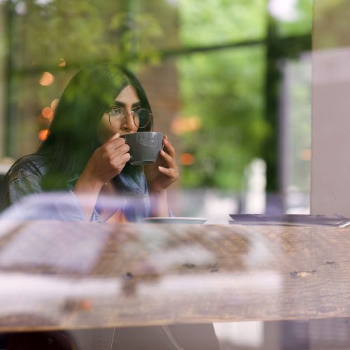 一位年轻的印度妇女戴着眼镜，坐在玻璃窗后面喝着咖啡。