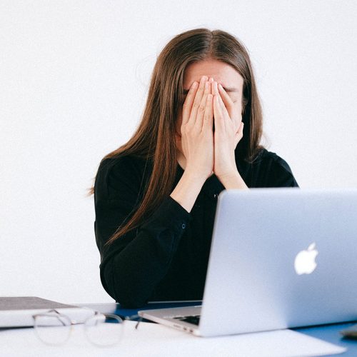 一位身穿黑色衬衫、留着棕色长发的女士坐在电脑前，双手抱着头。