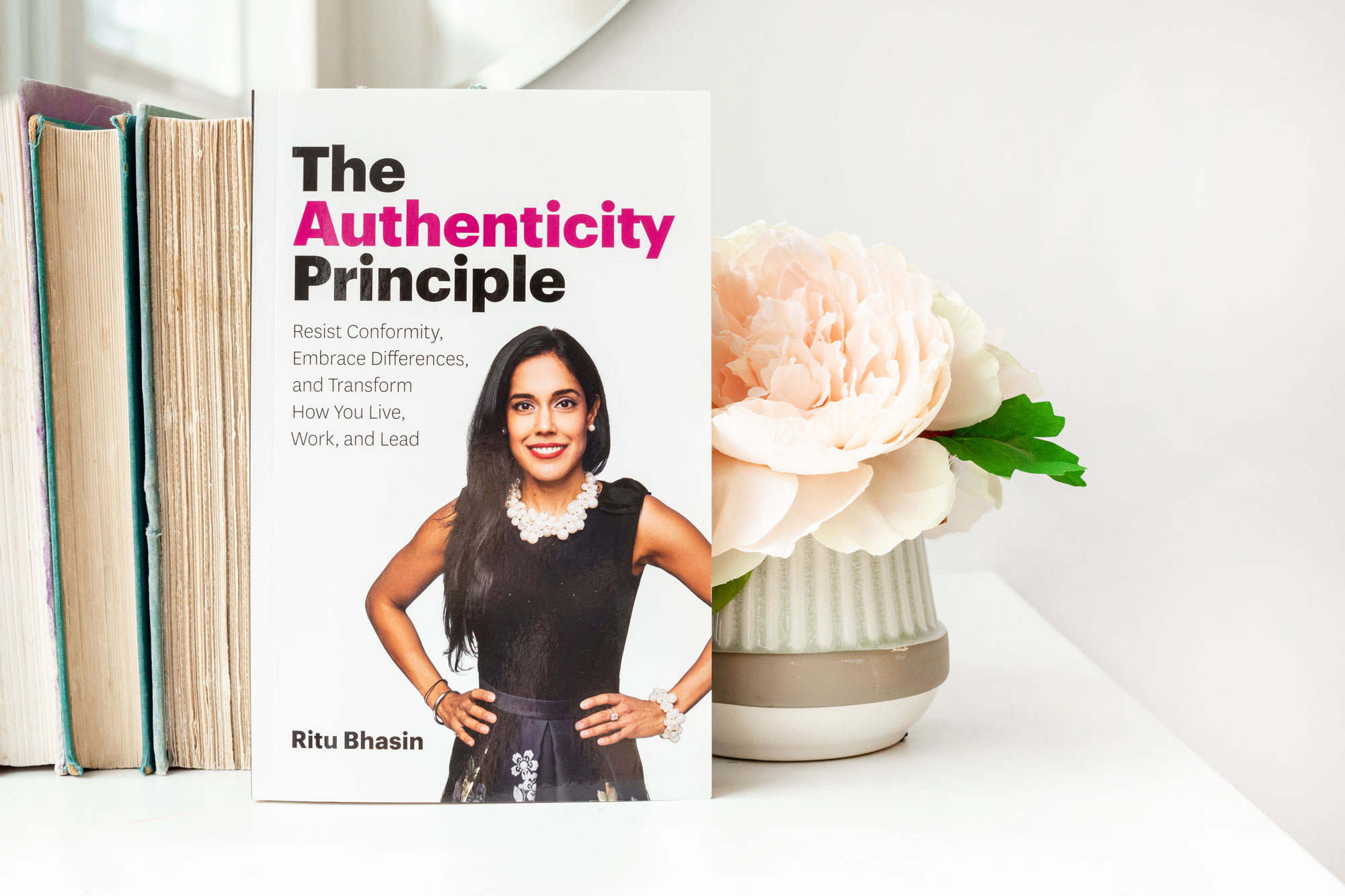 beplay体育手机里图·巴辛(Ritu Bhasin)的《真实性beplay最新备用网址原则》(The Authenticity Principle)坐在一张白色的桌子上，前面是一堆旧书，书页朝前，还有一个花瓶，上面插着一大朵淡粉色的花。