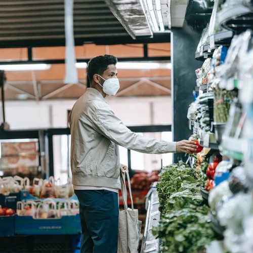 一个穿着奶油色外套，蓝色裤子和白色面罩的南亚男子从杂货店架子上捡起农产品