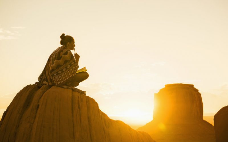 太阳升起时，一个女人披着有图案的披肩，拿着一本日记本坐在山上