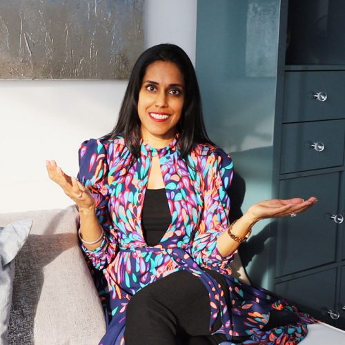 beplay体育手机Ritu Bhasin穿着彩色图案的连衣裙，坐在灰色的沙发上，微微抬起双手。