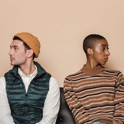 两个人并排坐在一张黑色的沙发上，互相望着对方。左边的人是白色的，穿着白色的衬衫和深绿色的羽绒背心，戴着黄色的帽子。右边的人是黑色的，留着寸头，穿着一件棕色、黑色和灰色相间的条纹毛衣。