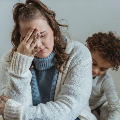 A young woman holding her head in her hand in exasperation with a small child behind her.