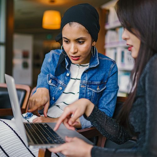 一名留着黑色长发、身穿黑色长袖上衣的亚洲女性和一名头戴黑色头巾、身穿蓝色牛仔夹克的棕色女性一起看着一台笔记本电脑，并指着屏幕。
