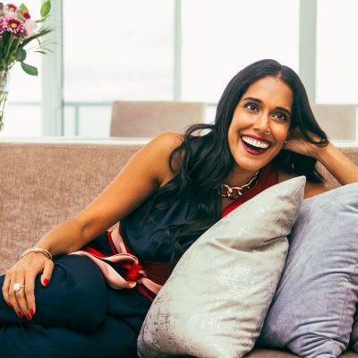 Ritu Bhasin sitting on a coach, leaning on three cushions with one hand on her folded legs and one hand in her hair. She is smiling authentically and looking off to her left.