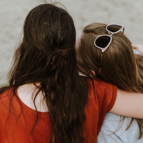 两名年轻妇女坐在海滩上的背景，一只胳膊围绕着另一个女人。