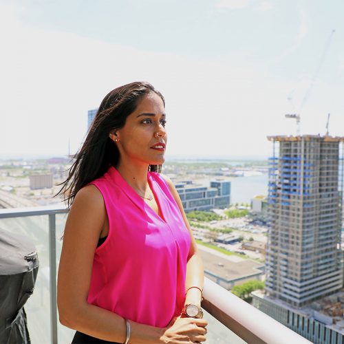 beplay体育手机Ritu Bhasin穿着一件亮粉色的无袖上衣，披头散发，站在公寓楼的阳台上，在阳光下向右侧张望