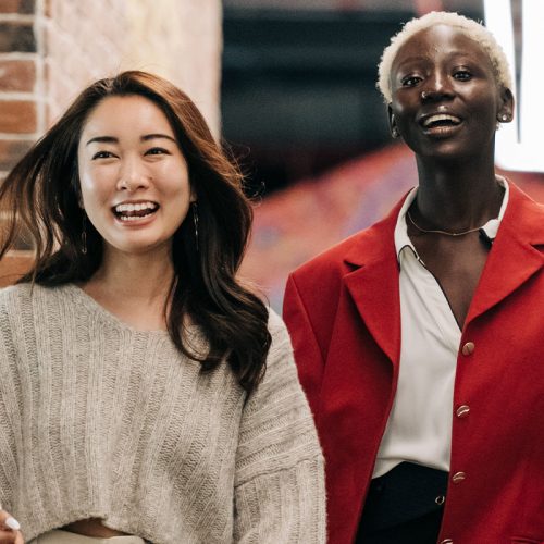 一个留着黑色长发的亚洲女人穿着米色毛衣，一个留着金色短发的黑人女人穿着红色外套，一边走一边微笑