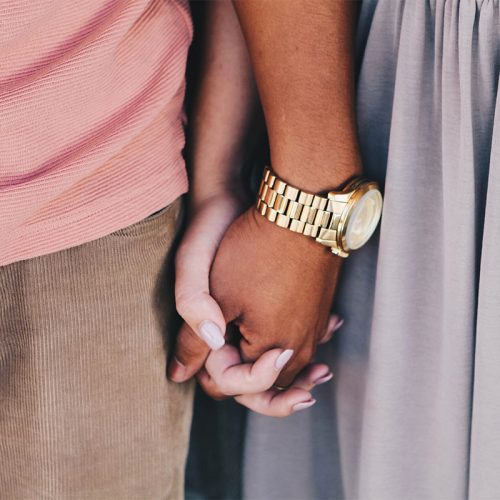 两个人手牵着手，一个深棕色皮肤的人戴着金色的手表，另一个肤色较浅的人涂着灰色的指甲油