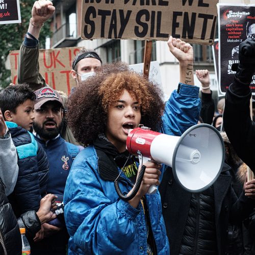 一名身穿蓝色夹克的年轻黑人妇女，右手举着扩音器，站在一群反种族主义抗议者旁边。